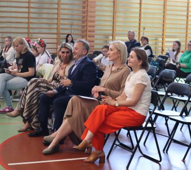 Oświetlona sala gimnastyczna. W pierwszym rzędzie na krzesłach siedzą elegancko ubrani dorośli. W tle pozostałe osoby oraz...