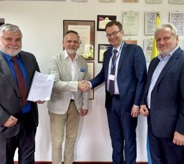 Wnętrze budynku. Czterech uśmiechniętych mężczyzn pozuje do zdjęcia. Dwóch z nich sciska sobie dłonie.