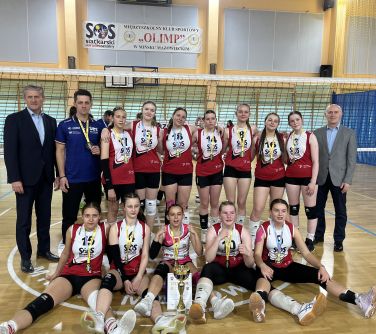 Wnętrze hali sportowej. Uśmiechnięte siatkarki w czerwonych koszulkach pozują do zdjecia w towarzystwie trzech mężczyzn.