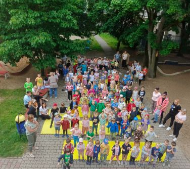Na dworze, plac, dzień. Grupa dzieci i opiekunów na chodniku. W tle drzewa. Zdjęcie z góry