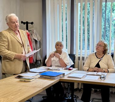 Oświetlone pomieszczenie. przed stołem przemawia mężczyzna. Na stołach dokumenty. Siedza dwie kobiety przed oknami. Za...