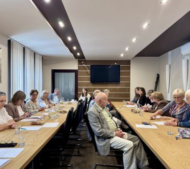 Oświetlona sala. Po obu stronach stoły. Przy nich osoby. Na stole dokumenty i dzbanki z woda
