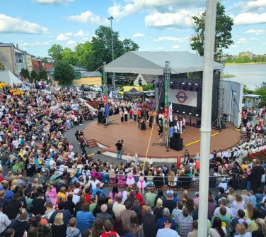 Słoneczny dzień. W amfiteatrze widownia. Na scenie wystepy. W tle jezioro