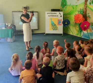 Oświetlona sala przedszkolna. Na niebieskim dywanie tyłem siedzi grupa dzieci. Na przodzie przed dzieśmi stoi kobieta