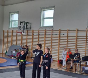 Hala sportowa. Na szarej macie trzy osoby. Kobieta w środku podnosi rękę dziewczynki z lewej. W tle drewniane drabinki