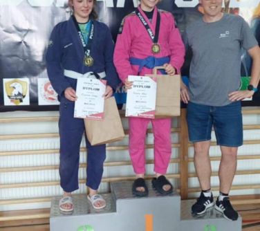Jasne pomieszczenie. na podium trzy osoby. Dwie kobiety i jeden mężczyzna. Kobiety z torbami i dyplomami
