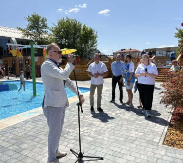 Sloneczny dizeń. Mężczyzna w jasnej marynarrce przemawia do mikrofonu. Za nim płytki basen. Obok pozostałe osoby