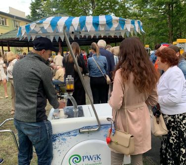 Na dworze, pochmurno. Stoisko PWiK z dystrybutorem wody. Kilka osób w kolejce. W tle plenerowe spotkanie
