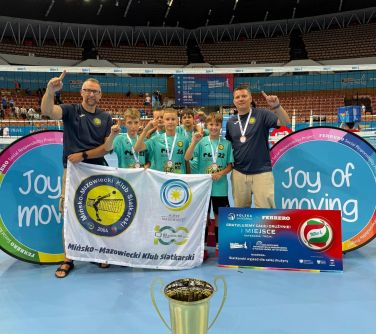 Drużyna pozuje z wielkim pucharem, medalami i banerem zwycięzców na tle hali Spodek. Po obu stronach chłopców trenerzy