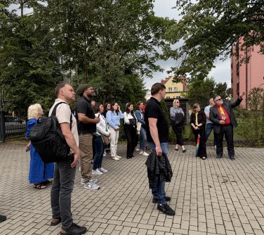 Pochmurny dzień. Zgromadzona grupa osób na chodniku słucha opowieści mężczyzny w ciemnym garniturze, czerwonej koszuli i...