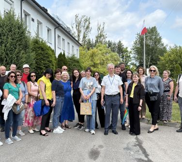 Dzień. Na chodniku grupa osób. W tle drzewa, biały budynek i maszt z flagą Polski