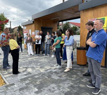Pochmurny dzień. Grupa ludzi stoi na chodniku z szarej kostki. Naokoło krzewy i drewniane wejście