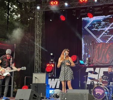 Dwór, scena. Kolorowe światla. Przy mikrofonie kobieta. po lewej mężcyznza z gitarą. Po prawej mężczyzna gra na perkusji