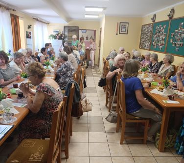 Oświetlone pomieszczenie. Po obu stronach stoły. Przy nich na krzesłach siedzą seniorzy