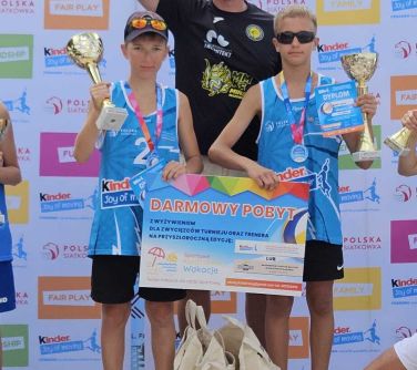 Dzień, na plaży. Na podium dwóch chlopców i za nimi mężczyzna który trzyma duży puchar. Chłopcy małe puchary i duży woucher