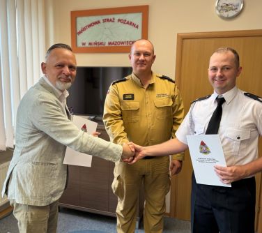 Jasne pomieszczenie. Trzech uśmiechniętych mężczyzn w uścisku dłoni. Dwoch z nich trzyma w dłoniach białe teczki