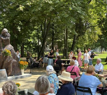 Słoneczny dzień. park. Po lewej pomnik obok w jasnym pojemniku żółte kwiaty. Po prawej na krzesłach siedzą osoby przy...