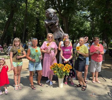 Słoneczny dzień. Park. Dziewięć kobiet z żółtymi kwiatkami pozuja przed pomnikiem kobiety. W tle drzewa