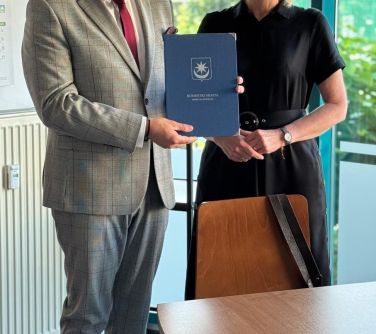 Jasne pomieszczenie. Uśmiechnięta kobieta i mężczyzna. Męzczyzna trzyma granatową teczkę w dłoni.