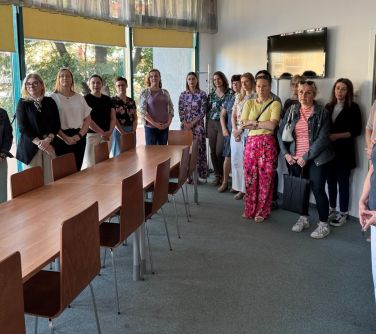 Jasne pomieszczenie. Grupa kobiet stoi w półkolu. Po środku długi stół z krzesłami