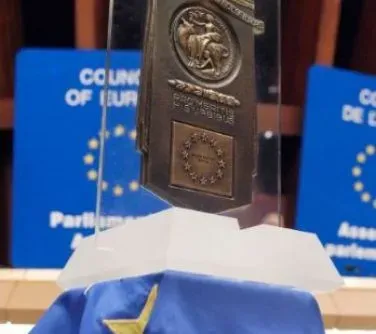 Medalion z symbolem Europarlamentu i gwiazdami Unii Europejskiej na niebieskiej fladze z gwiazdą, umieszczony na przezroczystym stojaku.
