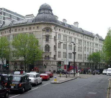 Duży zabytkowy budynek z kopulastym dachem widoczny przy ruchliwej ulicy z samochodami i drzewami.