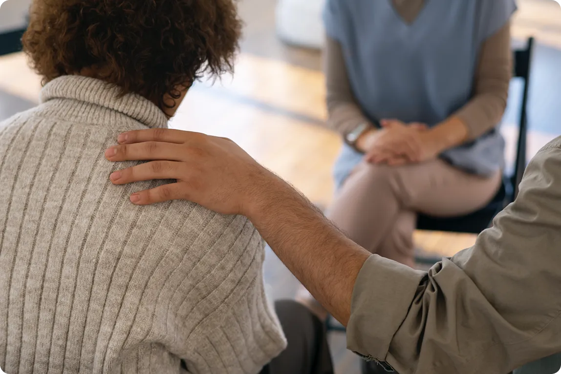 Ręka osoby dotykająca ramię drugiej osoby w szarym swetrze, z trzecią osobą siedzącą naprzeciwko.
