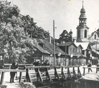 Kliknięcie powiększy obraz. Drewniany most z kilkoma przechodniami na tle drewnianych budynków i wieży kościoła.