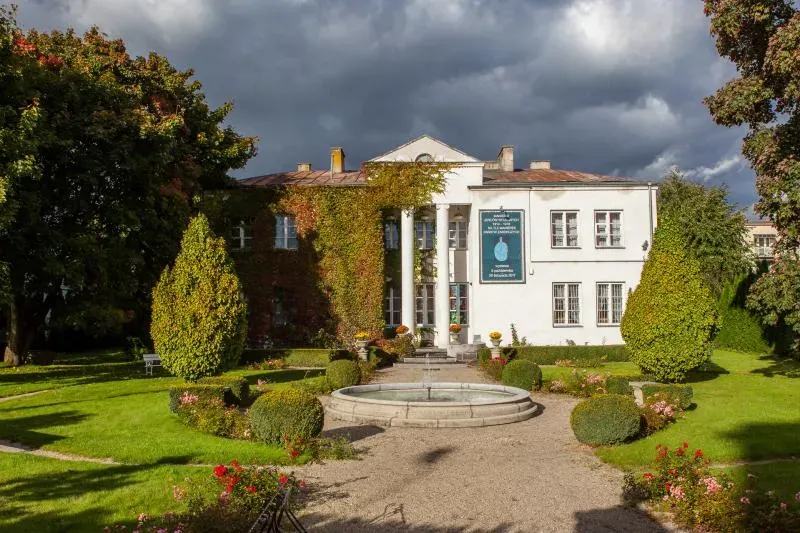 Biały budynek z dwiema kolumnami, otoczony ogrodem z fontanną i różnorodną roślinnością.