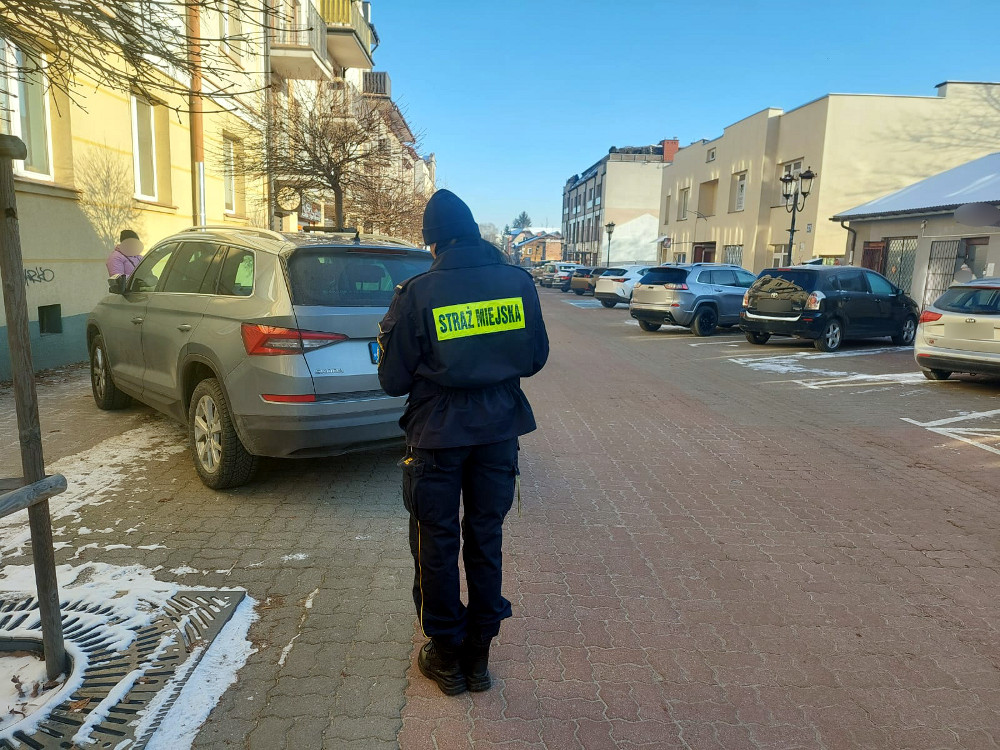 Na dworze w ciągu dnia, na dreptaku strażnik miejski tyłem, stoi za samochodem