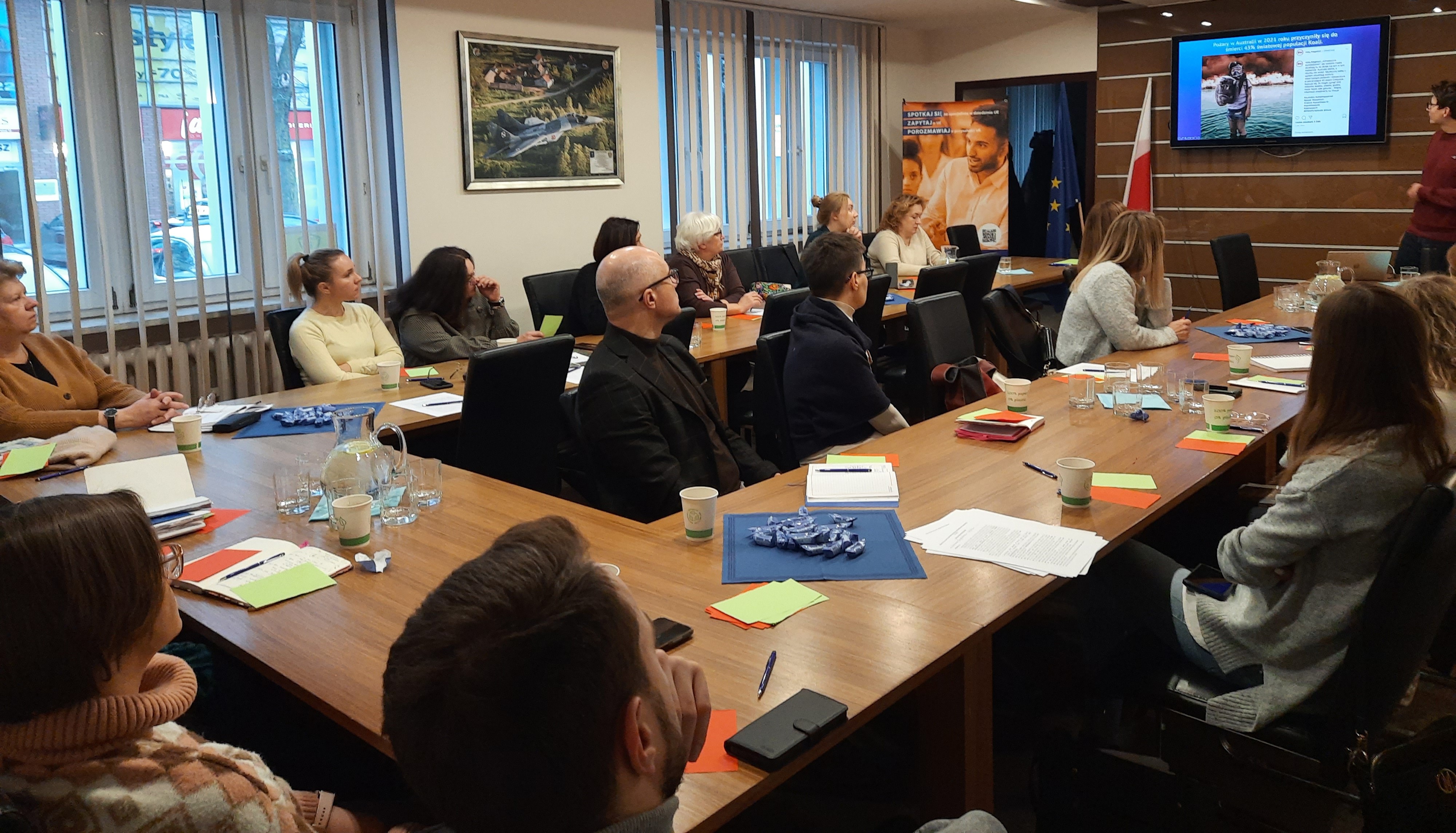 Sala konferencyjna. Na krzesłach przy stole ułożonym w podkowę siedzą osoby.