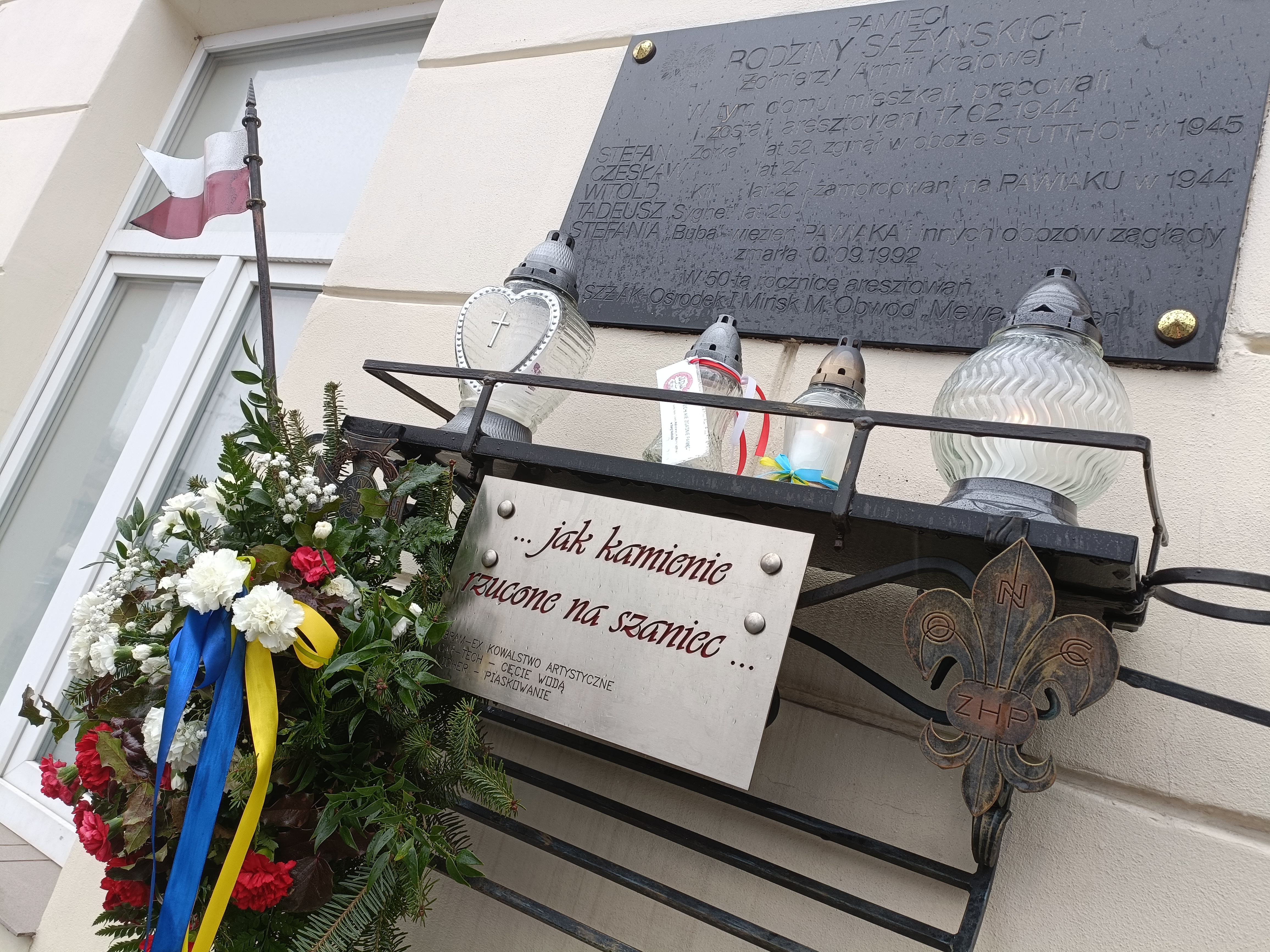 Na dworze, tablica upamiętniająca wisi na ścianie. Na zaczepionej pod tablicą półce stoją cztery znicze, na haczyku wisi wiązanka z biało-czerwonych kwiatów. Do półki zaczepiona tabliczka ...jak kamienie rzucone na szaniec...