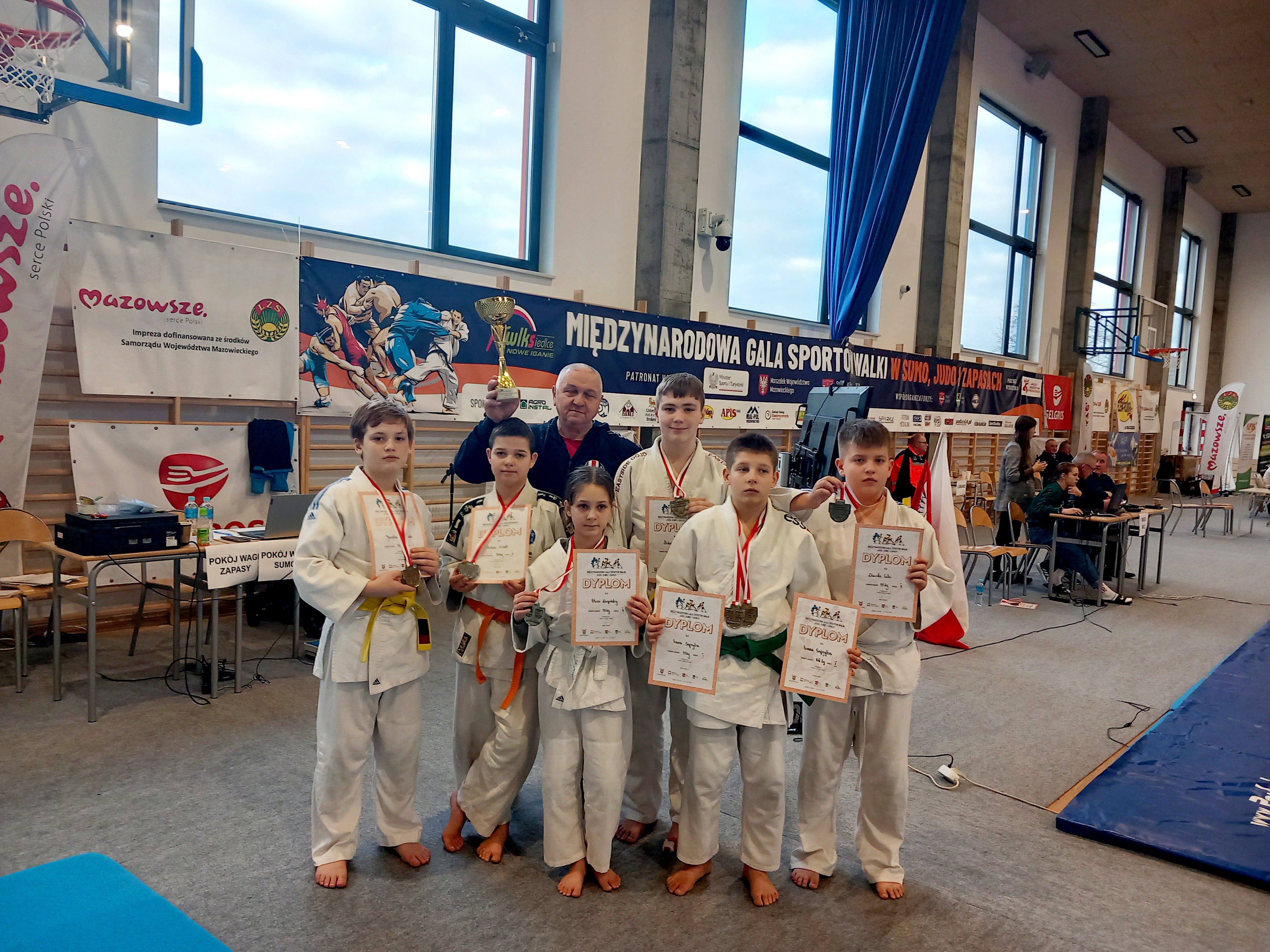 Hala sportowa. Sześcioro zawodników w kimonach. Trzymają w dłoniach dyplomy. Za nimi stoi mężczyzna z pucharem w dłoni.