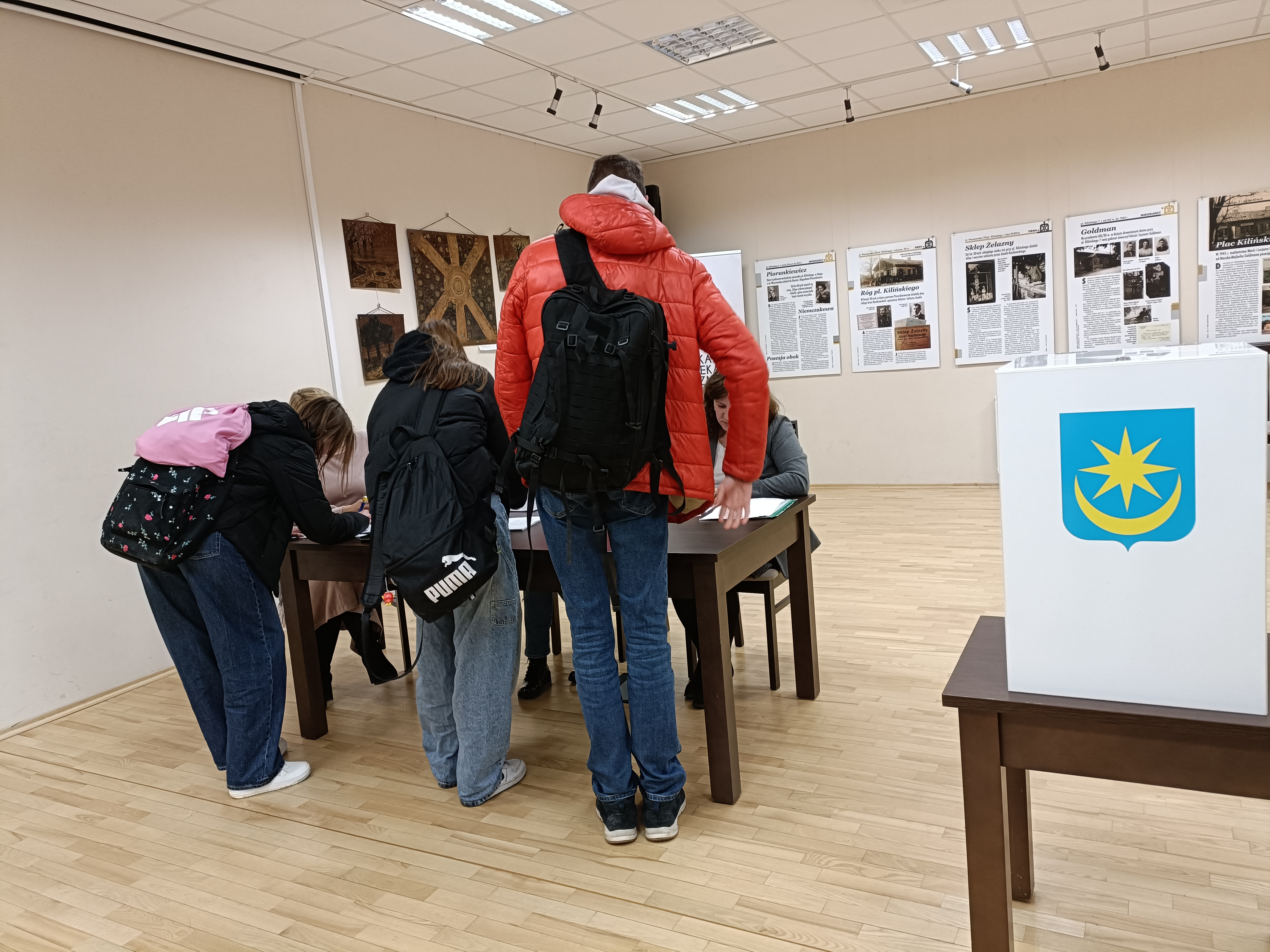 Pomieszczenie. Przed stołem trzech nastolatków w kurtkach. Pochylają się nad stołem. Podpisują dokumenty