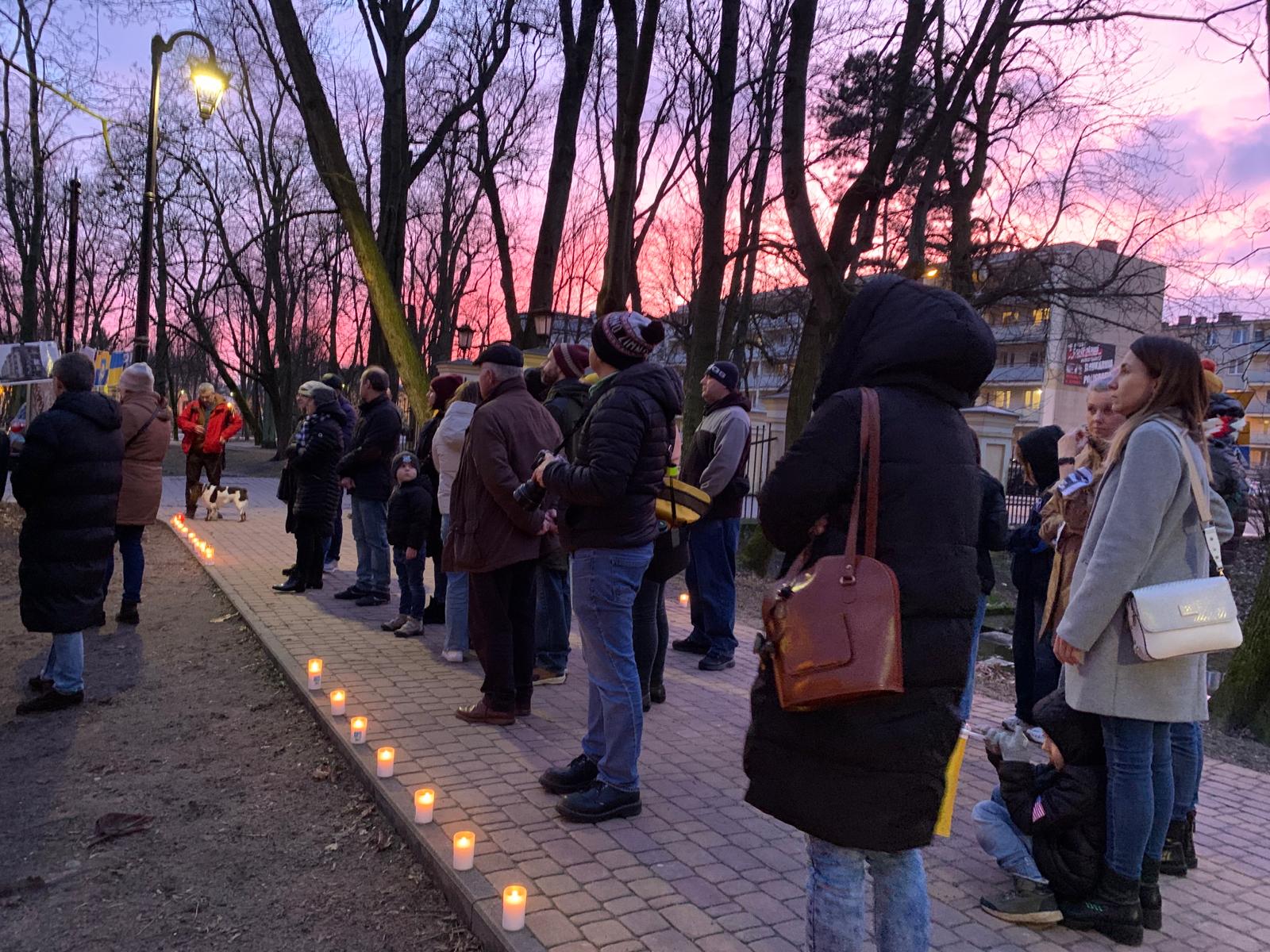 Alejka w parku wieczorową porą. Na niebie zachód słońca, niebo różowe. Na alejce z kostki stoją osoby. Wzdłóż alejki szpaler zapalonych zniczy.