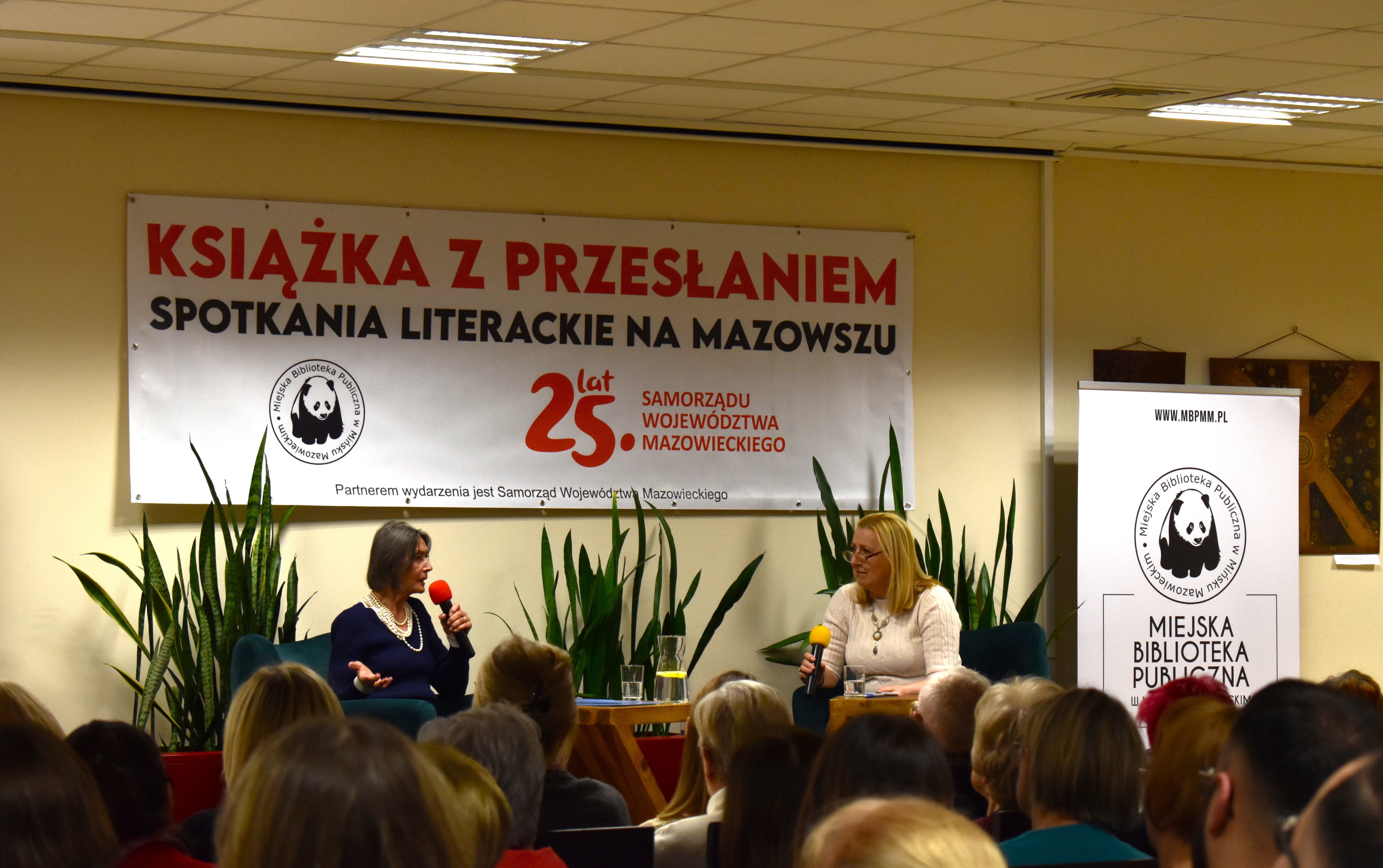Sala konferencyjna. Na dwóch fotelach siedzą kobiety. Po lewej kobieta przemawia do mikrofonu. Przed nimi stolik na nich dzbanek z wodą i szklanki. W tle na ścianie duży baner Książka z przesłaniem. Na przodzie na krzesłach słuchacze