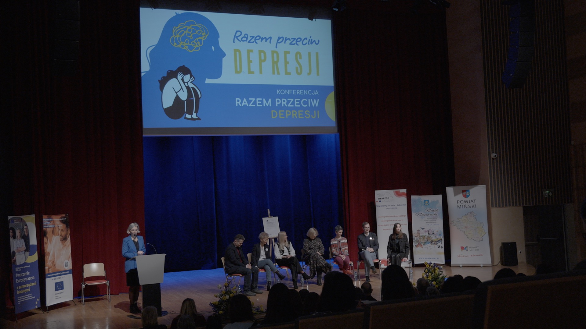 Scena. Przed mównicą przemawia kobieta. Na scenie po obu stronach roll-upy, na środku siedem osób siedzi na krzesłach. W tle zawieszony ekran z tytułem Razem przeciw depresji