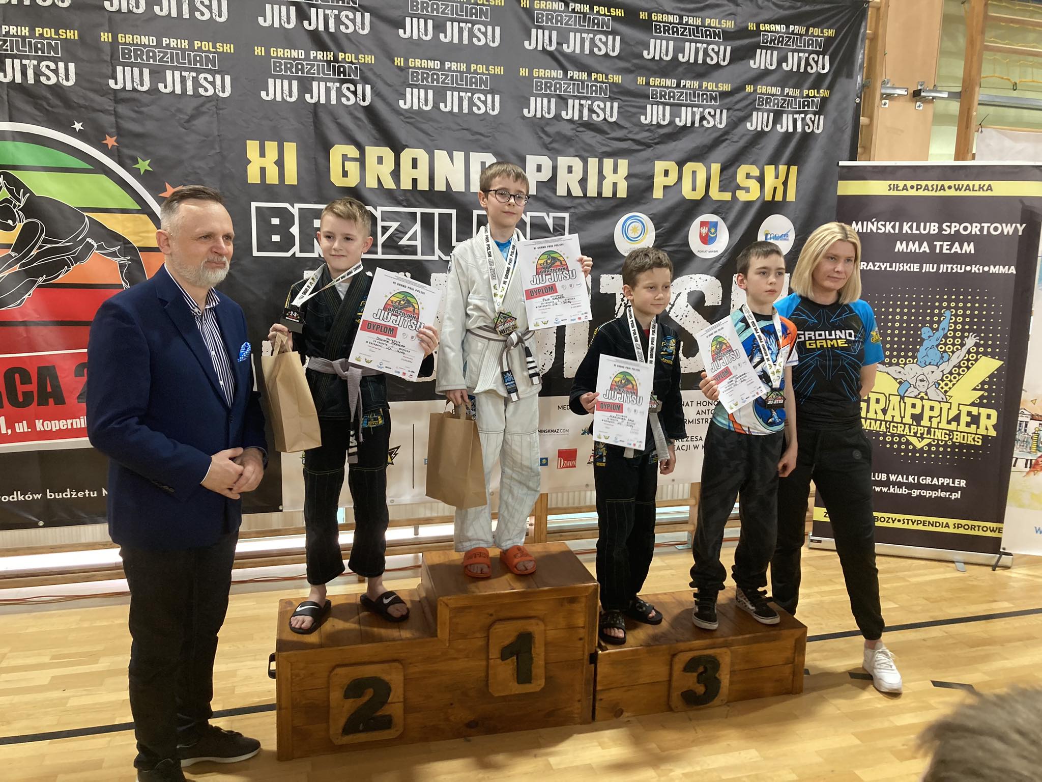 Hala sportowa. Dzieci pozują na podium z medalami i dyplomami w dłoniach. Po prawej kobieta w stroju sportowym. Po lewej mężczyzna w marynarce