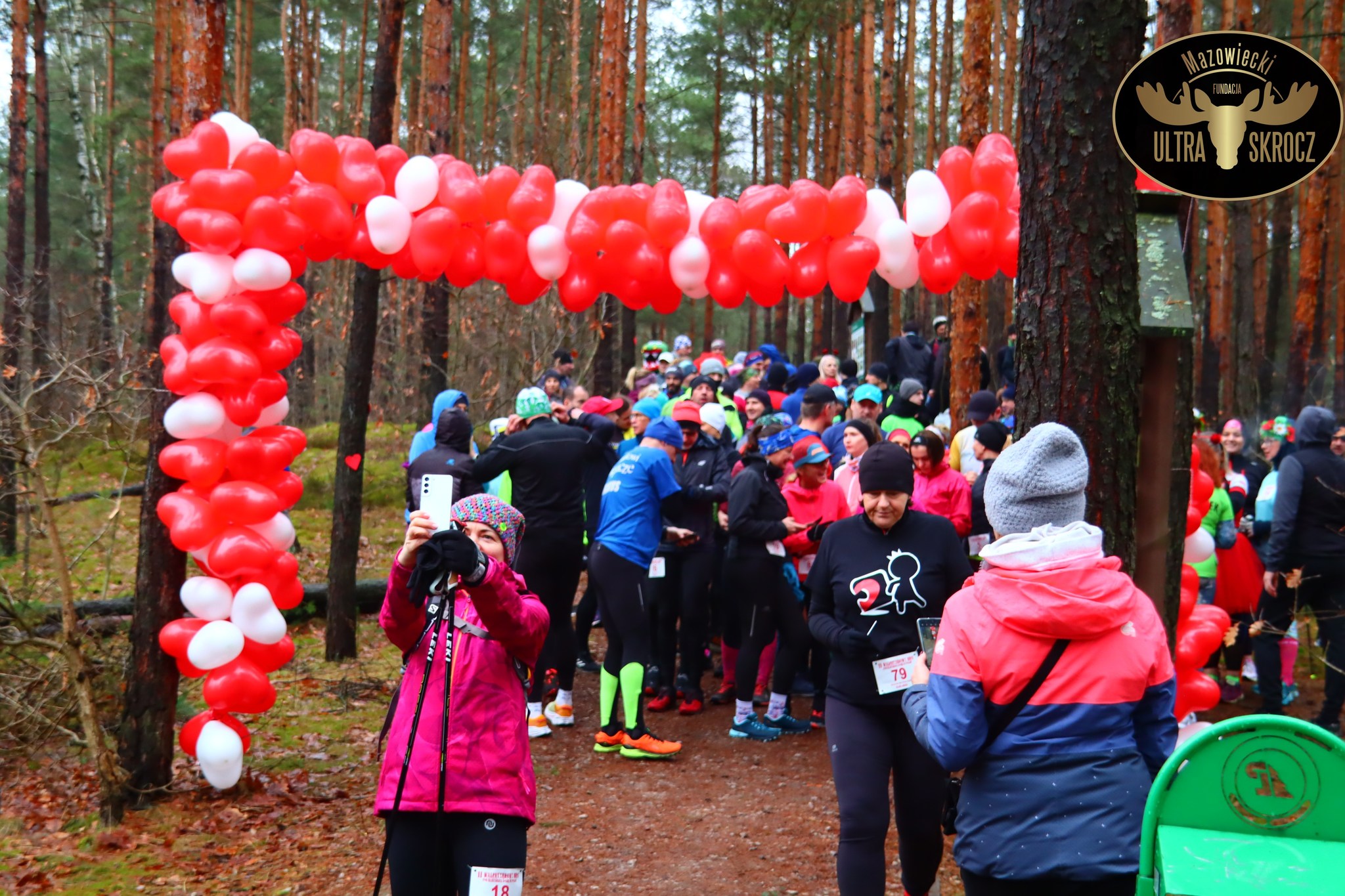 Ścieżka w lesie. Pomiędzy dzrzewami zawieszony łańcuch biało-czerwonych balonów wskazujący start biegu. Na starcie duża grupa osób