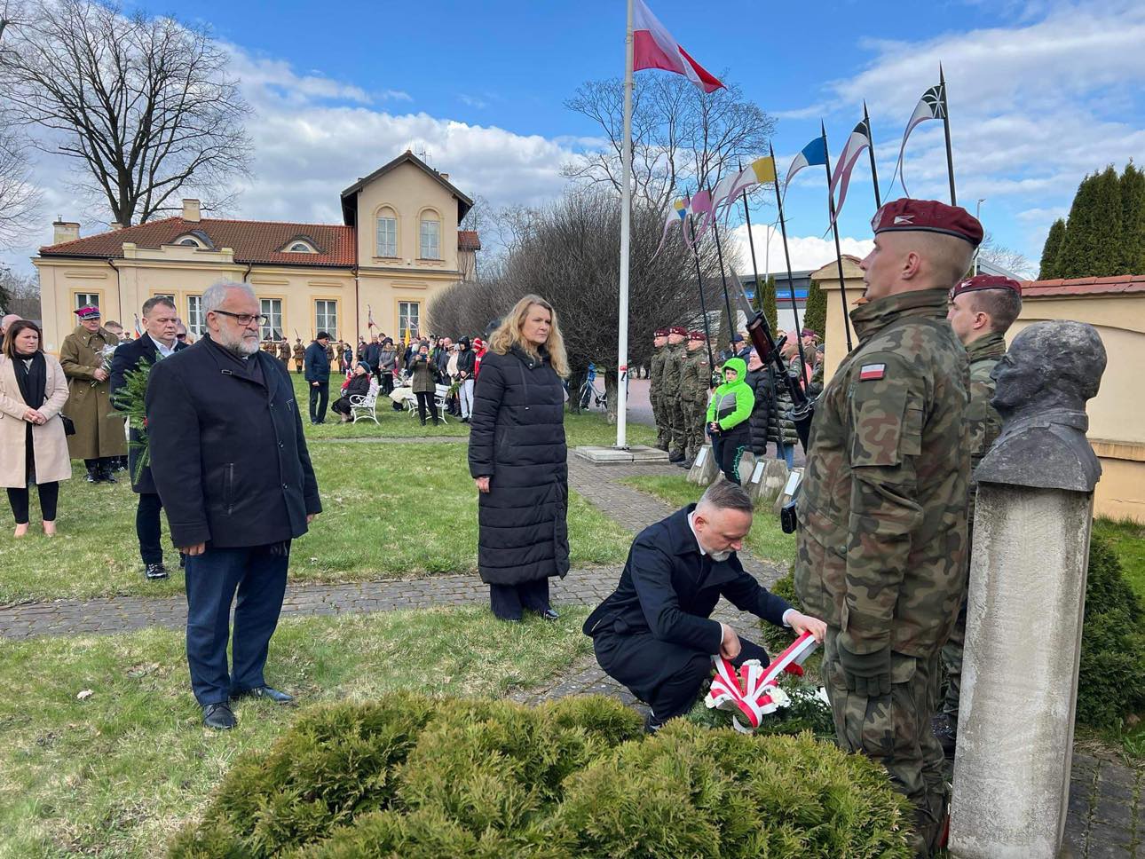 Na dworze dwóch mężczyzn i kobieta składają hołd przed pomnikiem. W tle osoby, żołnierze i budynek muzeum