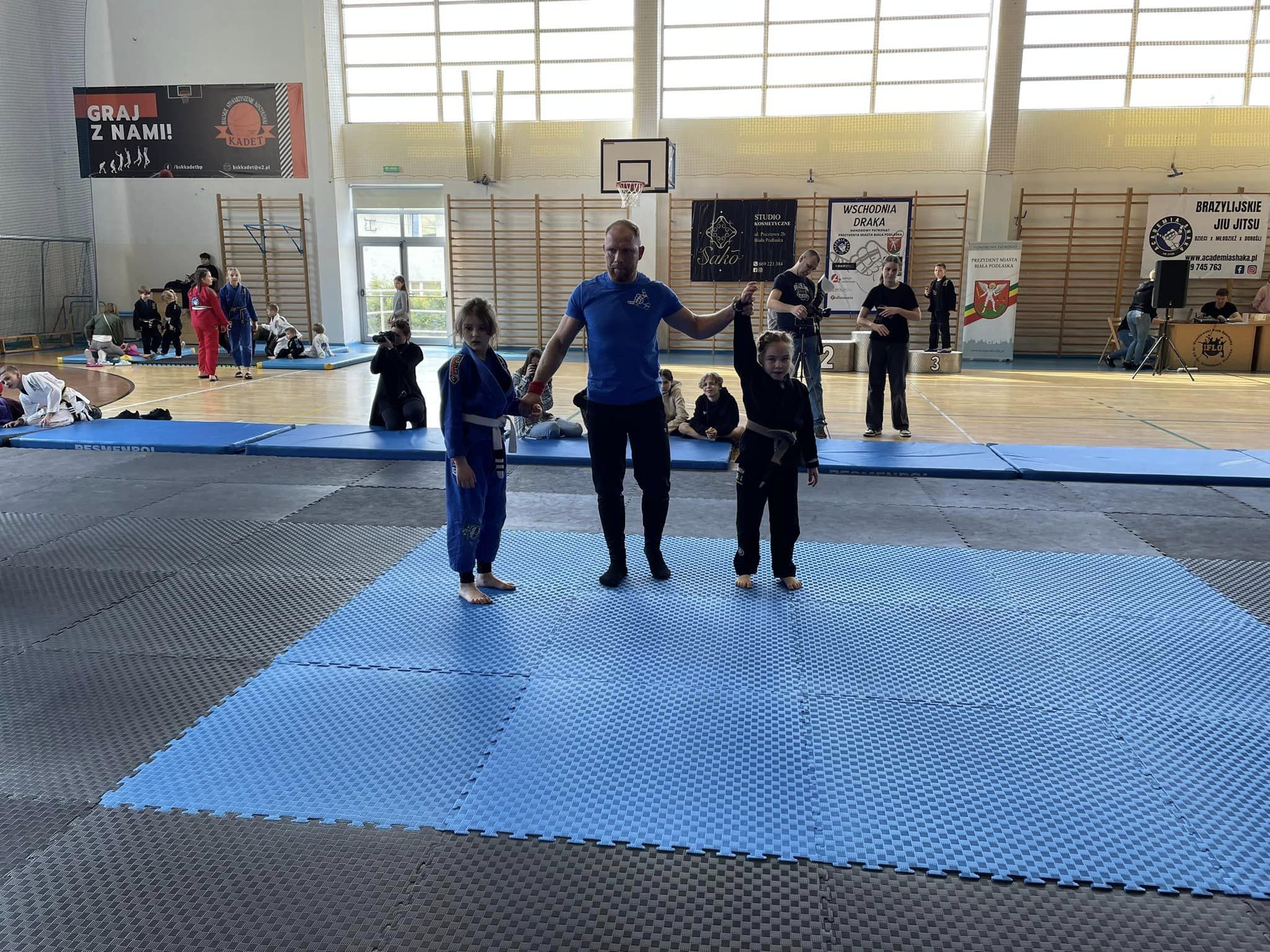 dziewczynki w kimonach stoją na macie sportowej, pomiędzy nimi stoi mężczyzna, unosi rękę jednej z nich do góry.