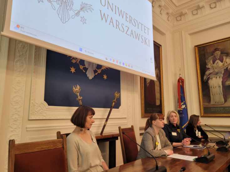 Pomieszczenie. Przy drewnianym stole siedzą cztery kobiety. Przed nimi mikrofony. Za kobietami na ścianie wisi ekran.