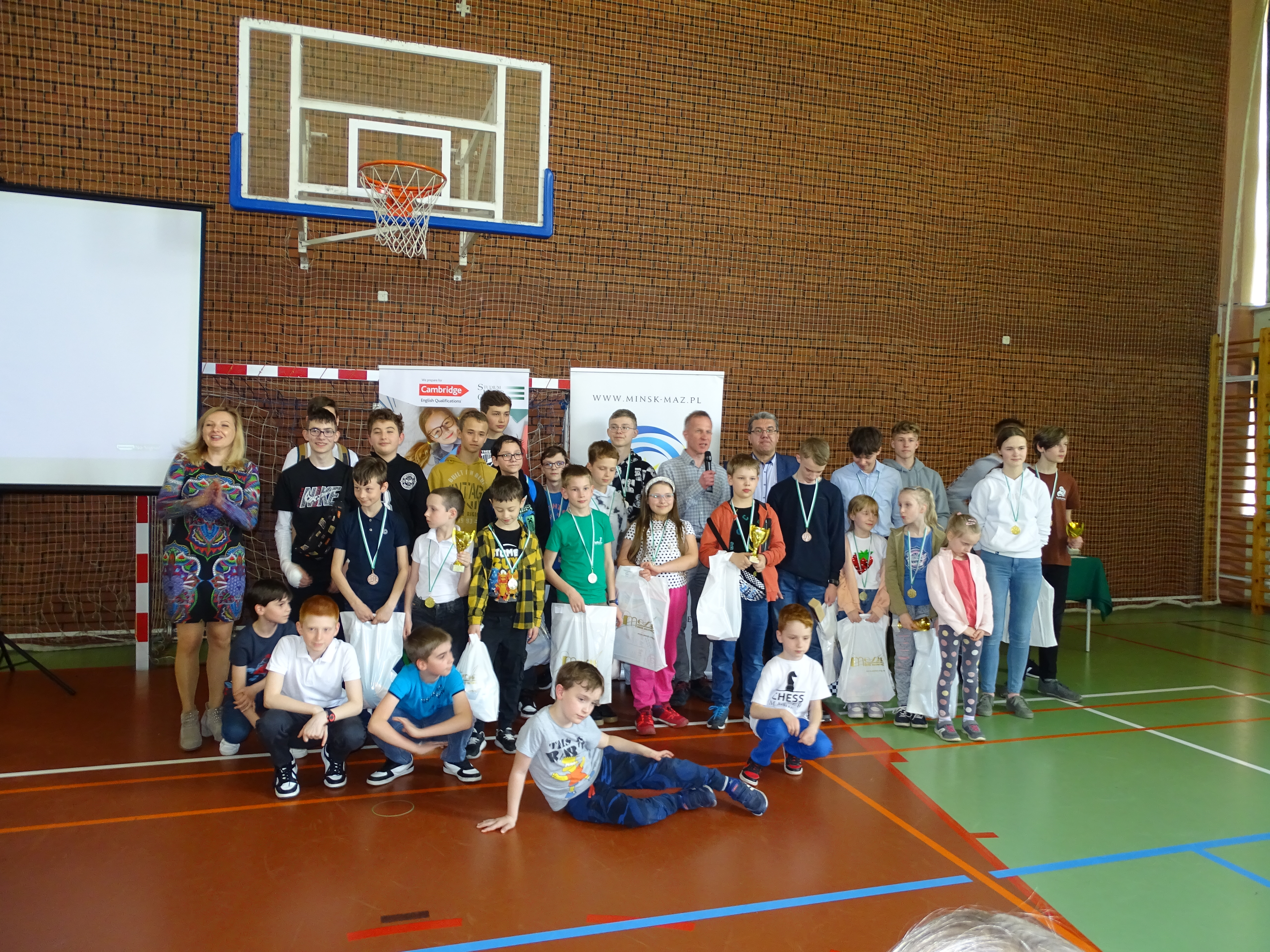 Hala sportowa. Grupa dorosłych oraz dzieci pozuje do zdjęcia, na środku mężczyzna mówi do mikrofonu.