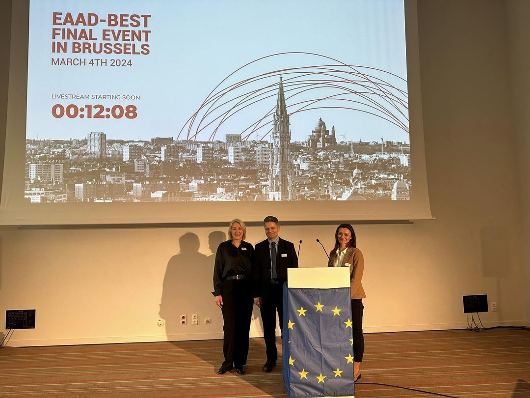 Aula. Trzy osoby stoja przed mównicą okalana flagą unii europejskiej. W tle wyświetlane zdjęcie miasta