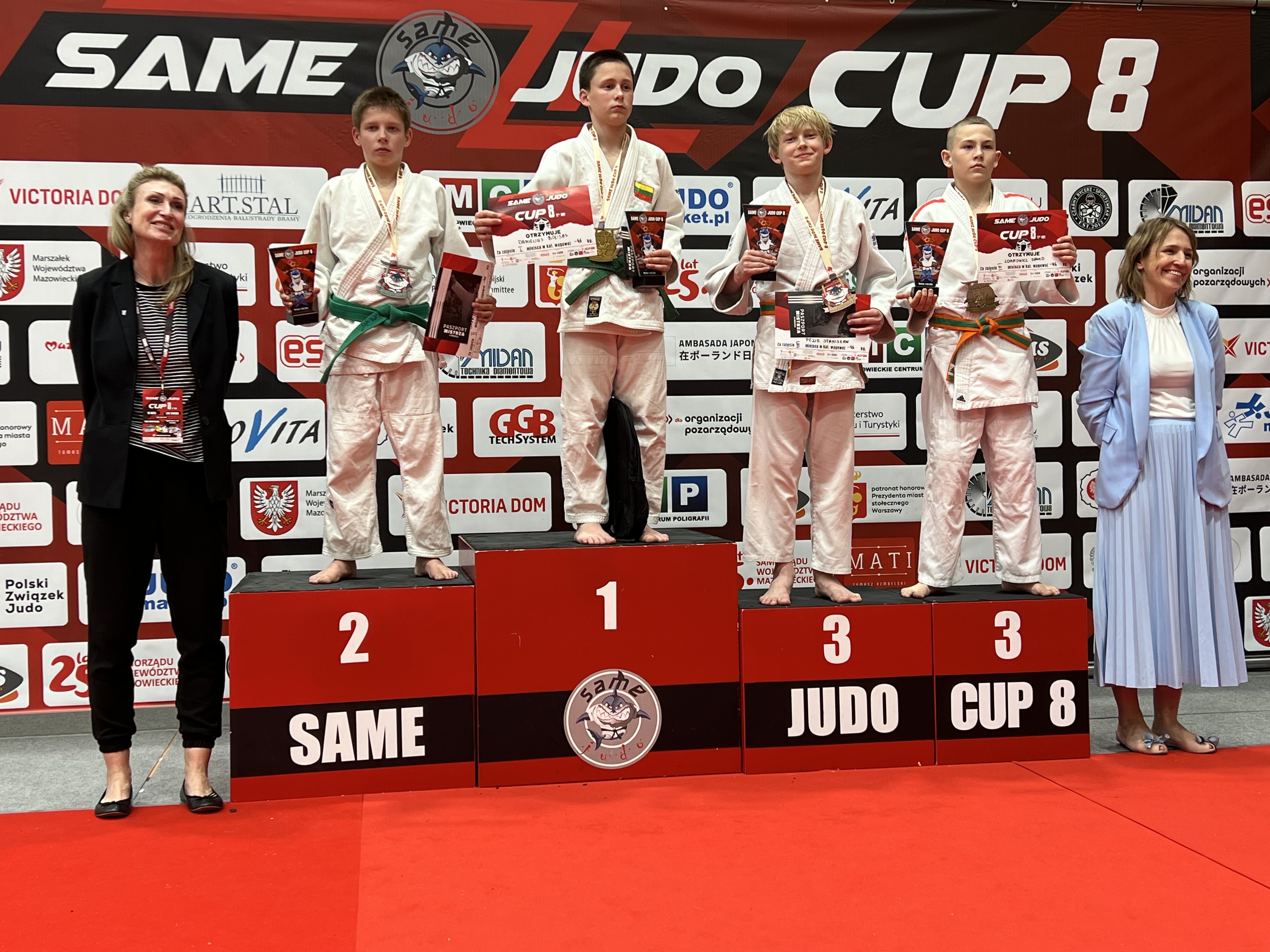 Hala. Na podium czterech chłopców w białych kimonach, dwóch na miejscu trzecim. Nagrody w dłoniach. Po obu stronach usmiechnięte kobiety