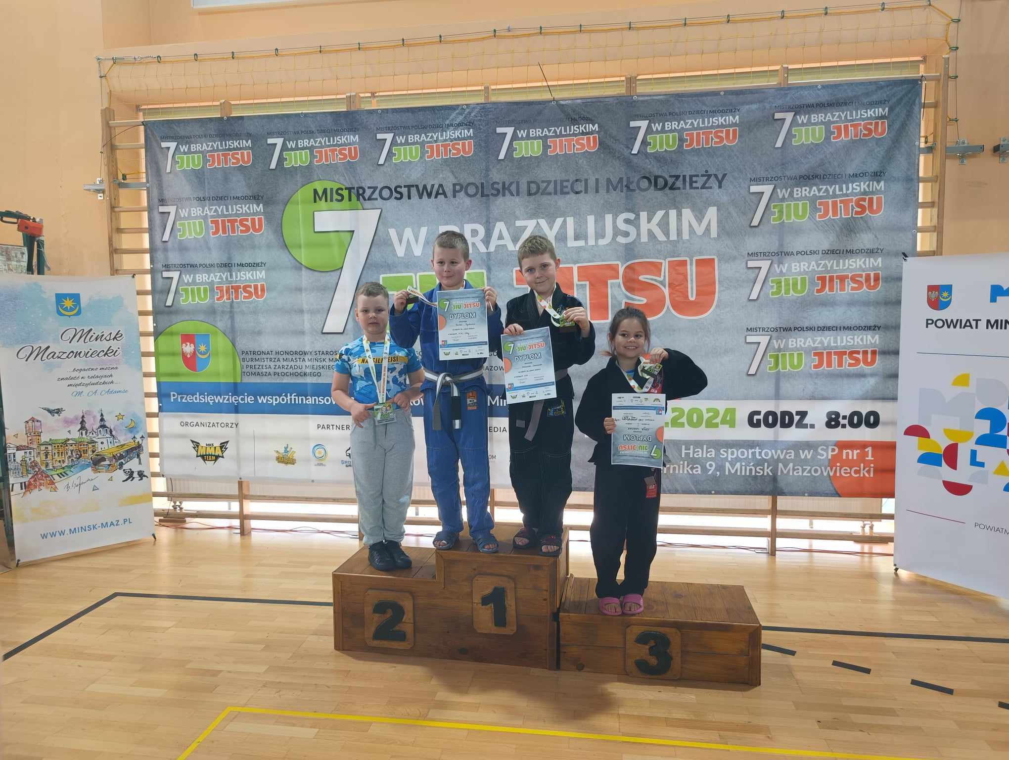 Hala sportowa. Czwórka dzieci z nagrodami w rękach stoją na podium. Dwie na miejscu pierwszym. W tle baner na drabinkach