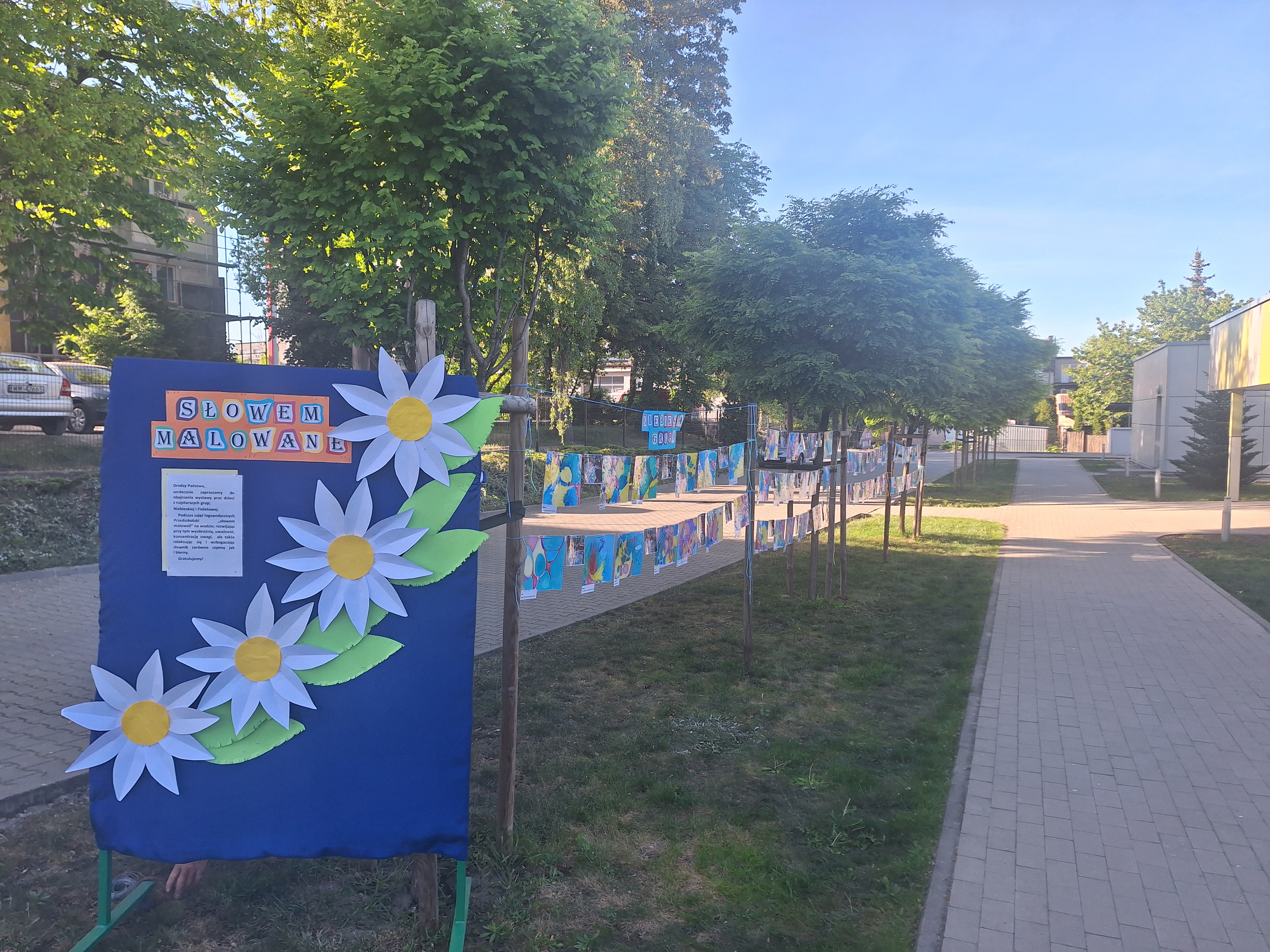 Dwór. Wzdłuż chodnika, na trawie, pomiędzy drzewami, na sznurkach rozwieszone kolorowe prace dzieci.