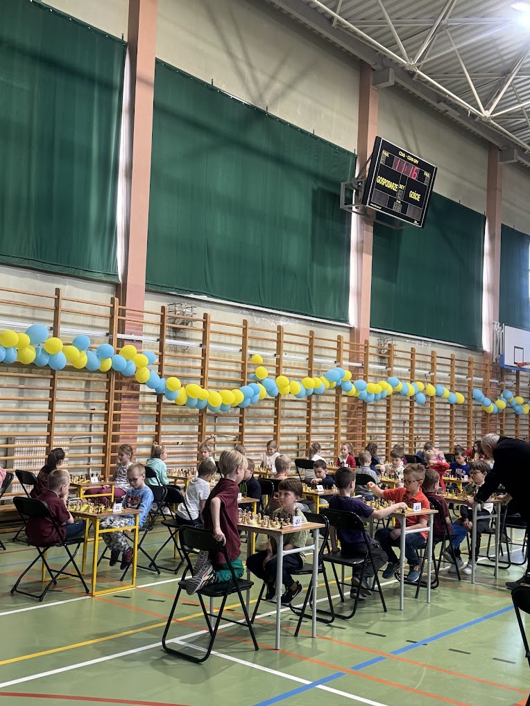 Sala gimnastyczna. Na stołach szachownice. Przy stołach dzieci rozgrywają parie. Po lewej żółto-niebieskie balony