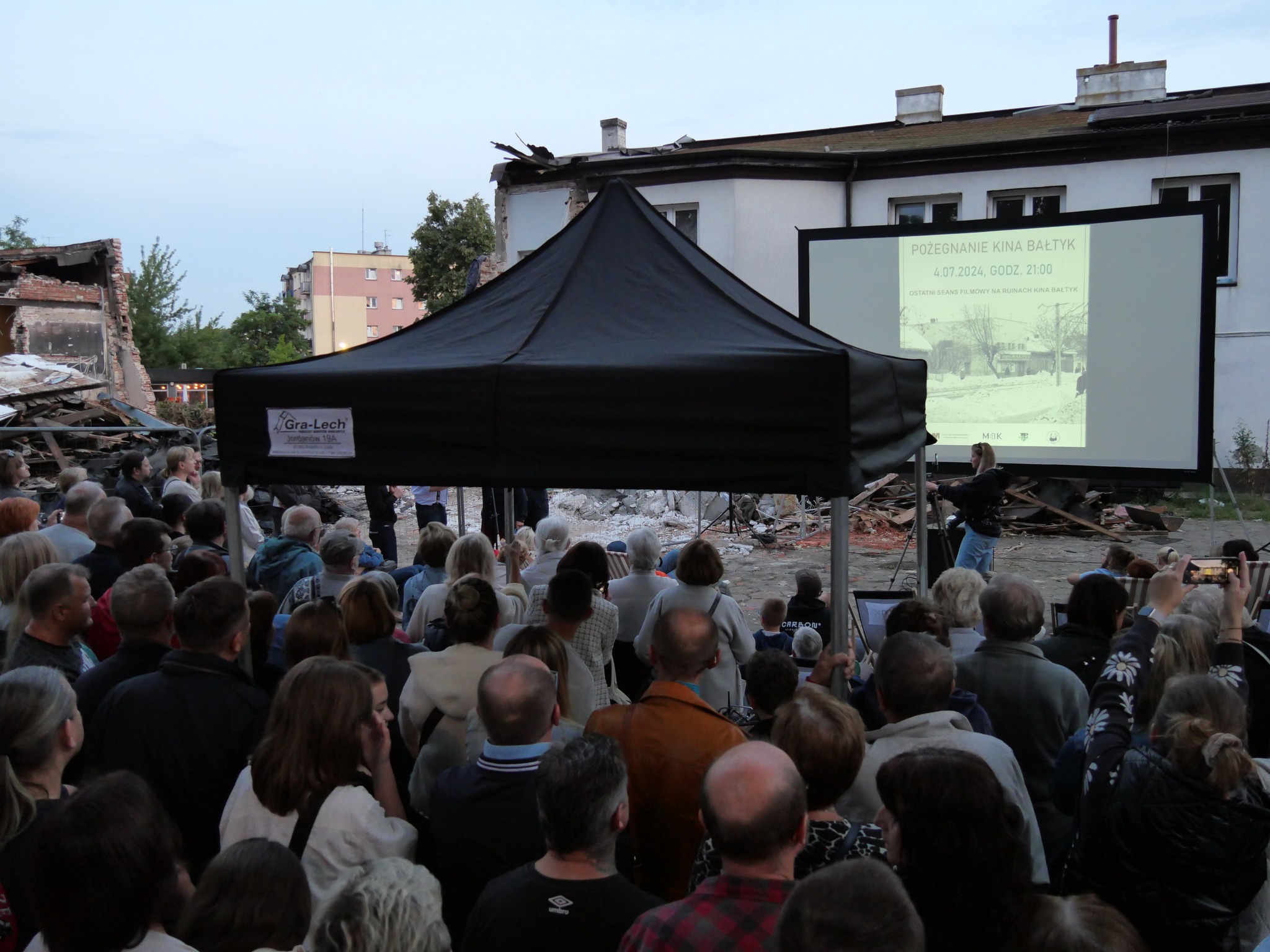 Za dnia duża grupa osób ogląda film wyświetlany na dużym ekranie. Po środku czarny namiot