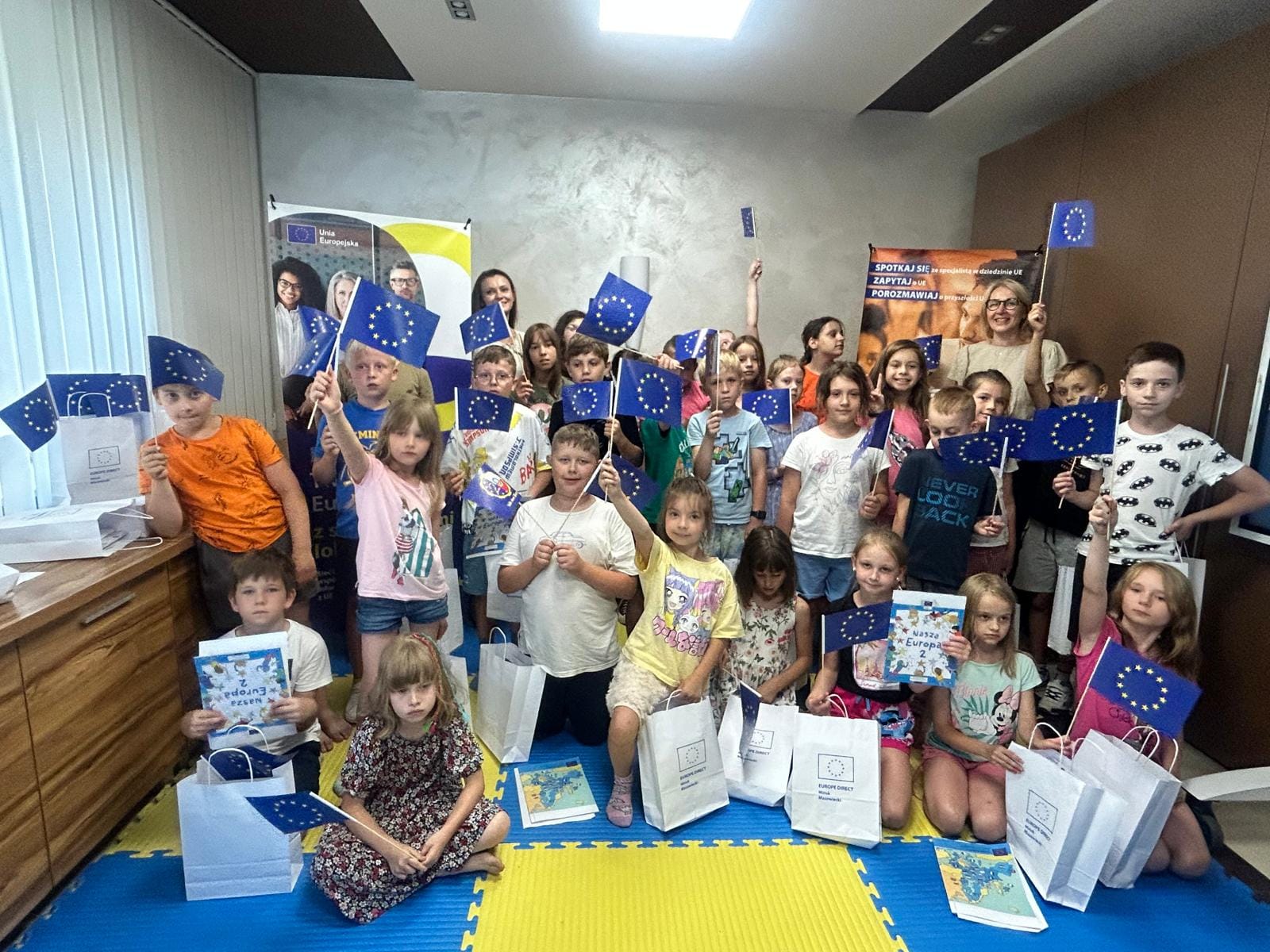 Jasne pomieszczenie. Grupa dzieci z flagami unii europejskiej. pozuja stojąc, siedząc na piankowych matach